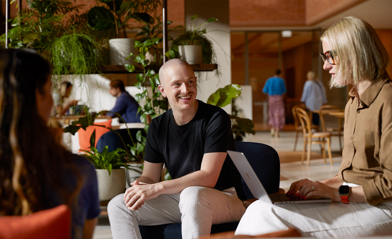 Troje pracowników Apple. Siedzą w fotelach w Caffè Macs i rozmawiają ze sobą. Jedna osoba ma na kolanach MacBooka.