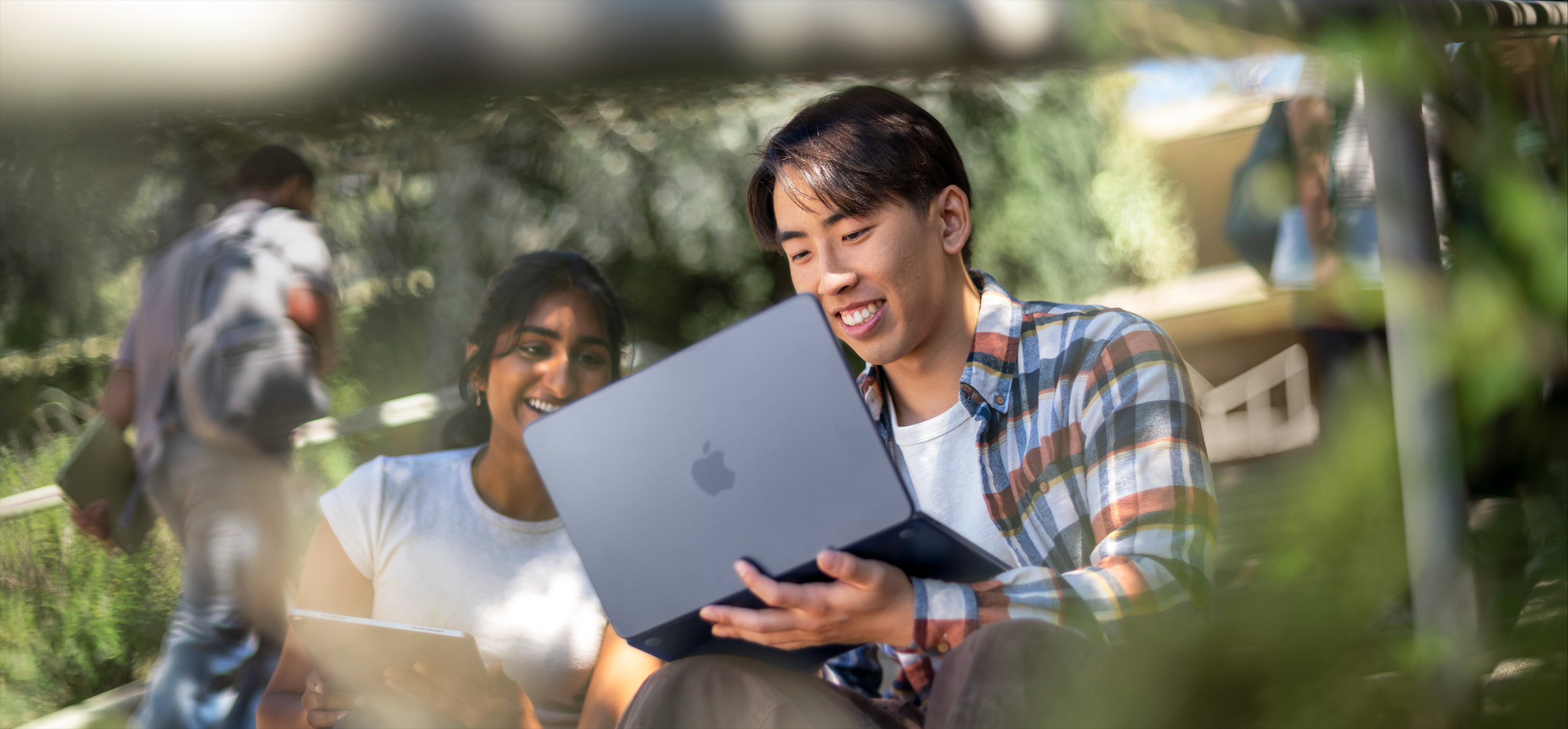 Due studenti sorridenti usano MacBook all’aperto, uno lo tiene sulle ginocchia mentre collaborano a un progetto