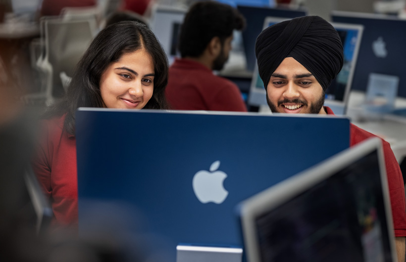 Dua mahasiswa yang tersenyum di dalam kelas melihat ke layar iMac yang mereka gunakan bersama.