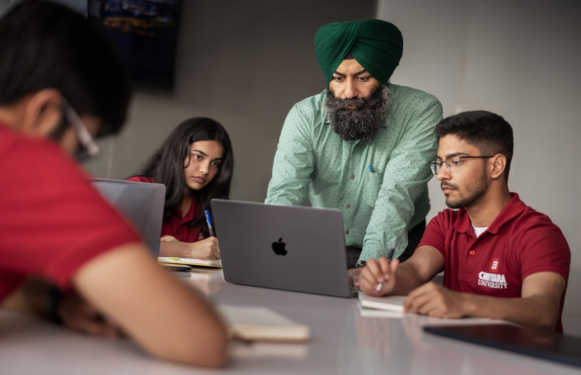 Seorang profesor di meja kelas membungkuk untuk melihat layar MacBook mahasiswanya.