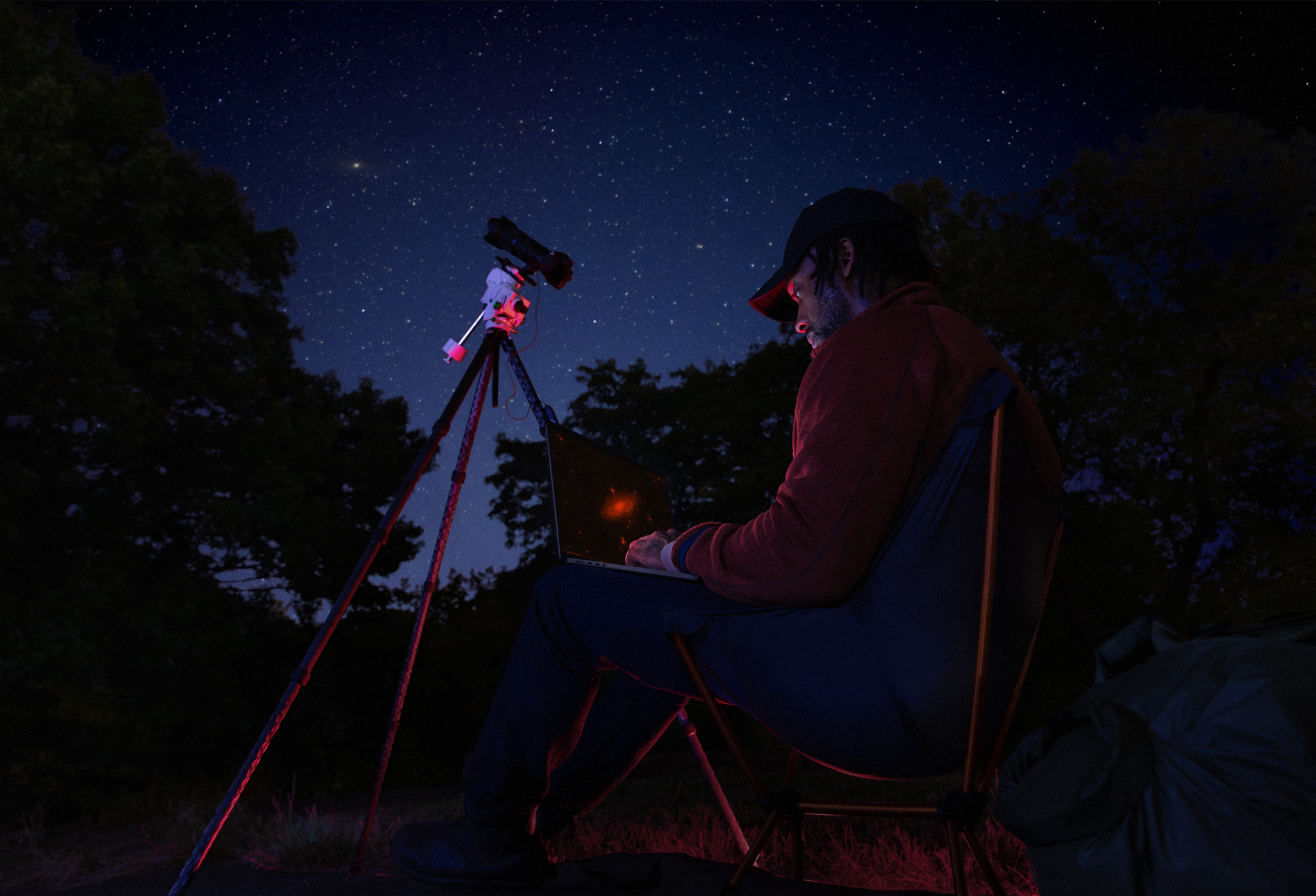 夜幕下，有個人坐在户外，將 MacBook Pro 放在腿上使用，MacBook Pro 連接了三腳架上一部對著星空的裝置